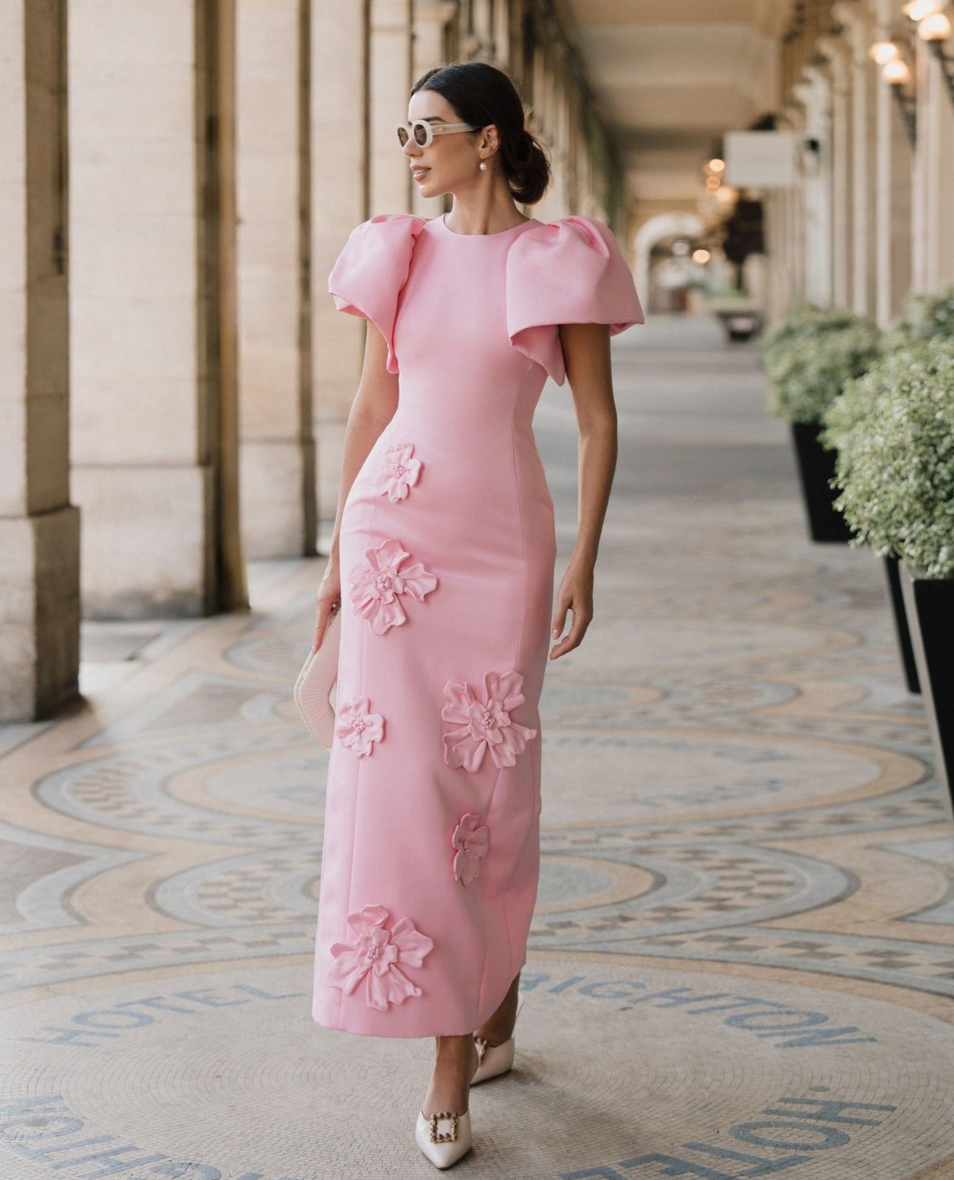 Woman in a pink dress with floral details walking along a decorative corridor.