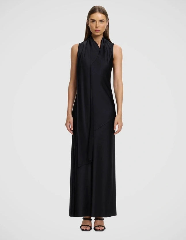 A woman modeling a sleeveless black wrap-style maxi dress with a high draped neckline and flowing asymmetrical skirt panels, photographed against a light gray studio background.